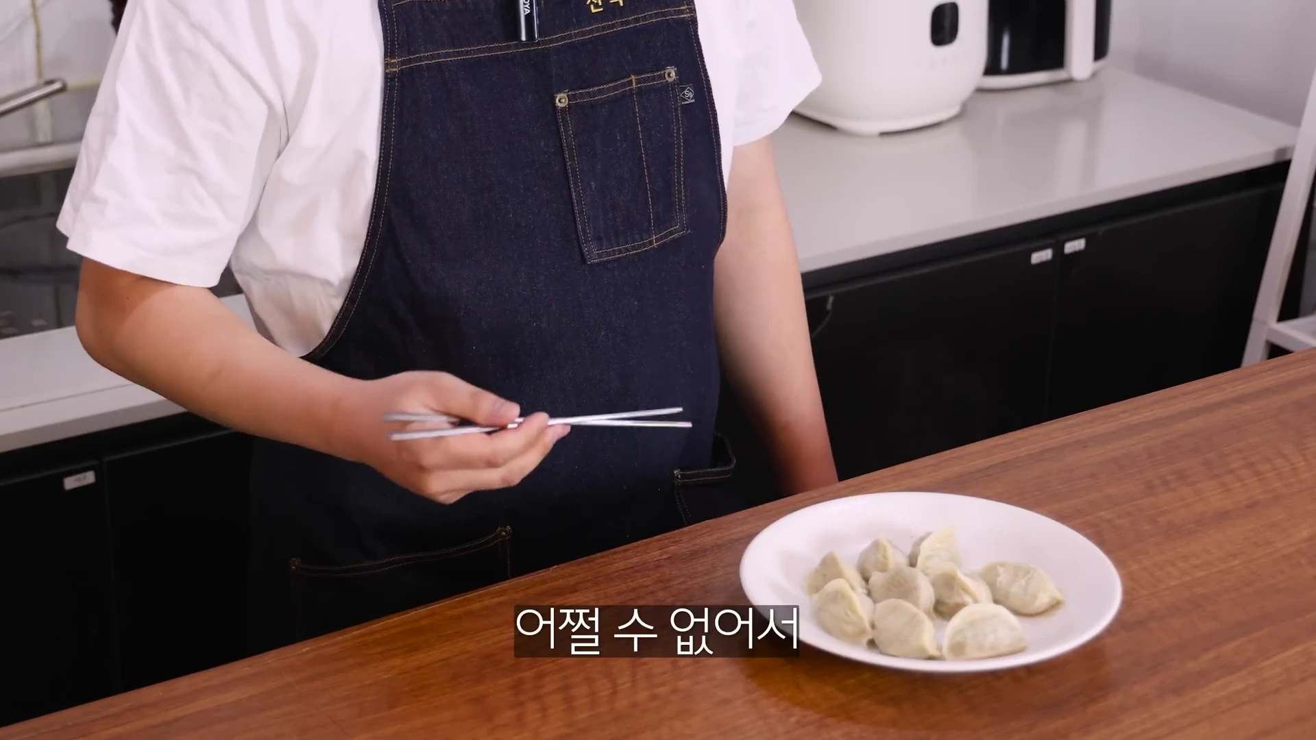 앞치마를 두른 요리사가 젓가락을 들고 접시에 담긴 만두를 가리키며 설명하는 모습