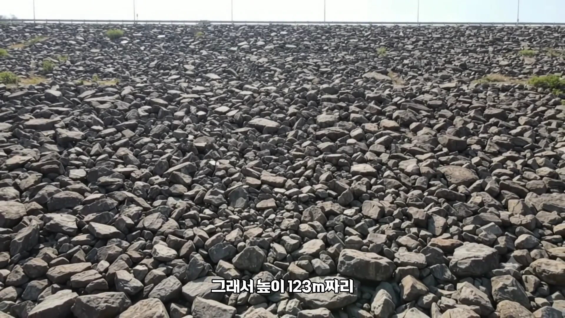 흙과 돌이 촘촘하게 쌓여 있는 거대한 사력댐의 경사면을 찍은 사진으로, 화면 하단에는 한국어 자막이 자막이 깔려 있습니다.