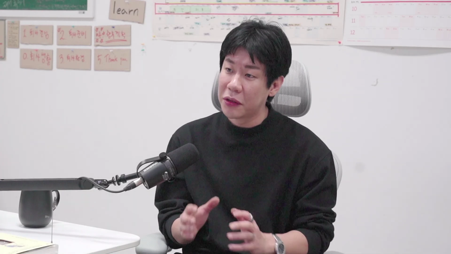 검은색 니트를 입은 남성이 마이크 앞에서 대화하며 손동작을 하고 있다. 뒤쪽 벽에는 'learn'이라는 글자와 숫자가 적힌 메모들이 부착되어 있다.