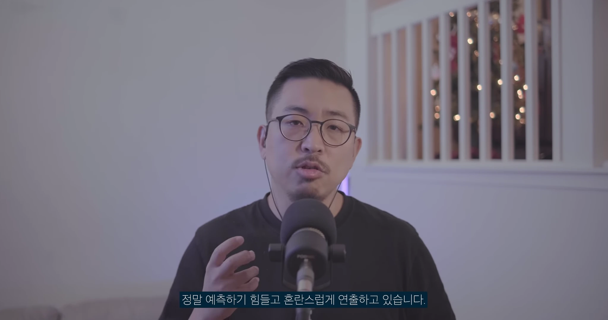 검은색 반팔 티셔츠를 입고 안경을 쓴 남성이 마이크 앞에서 손동작을 하며 이야기를 하고 있는 스튜디오 모습입니다.