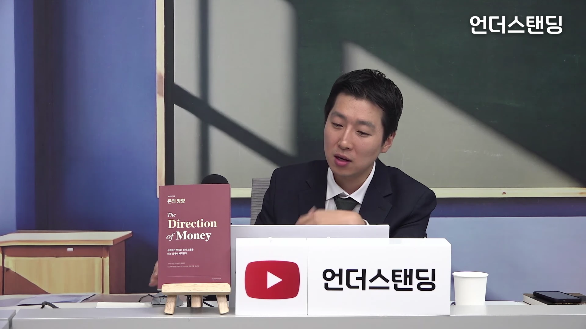 정장을 입은 남성이 스튜디오 테이블 뒤에 앉아 대화하며, 테이블 위에는 '돈의 방향'이라는 제목의 책이 스탠드에 놓여 있고 옆에는 채널 로고가 적힌 흰색 박스가 있음.