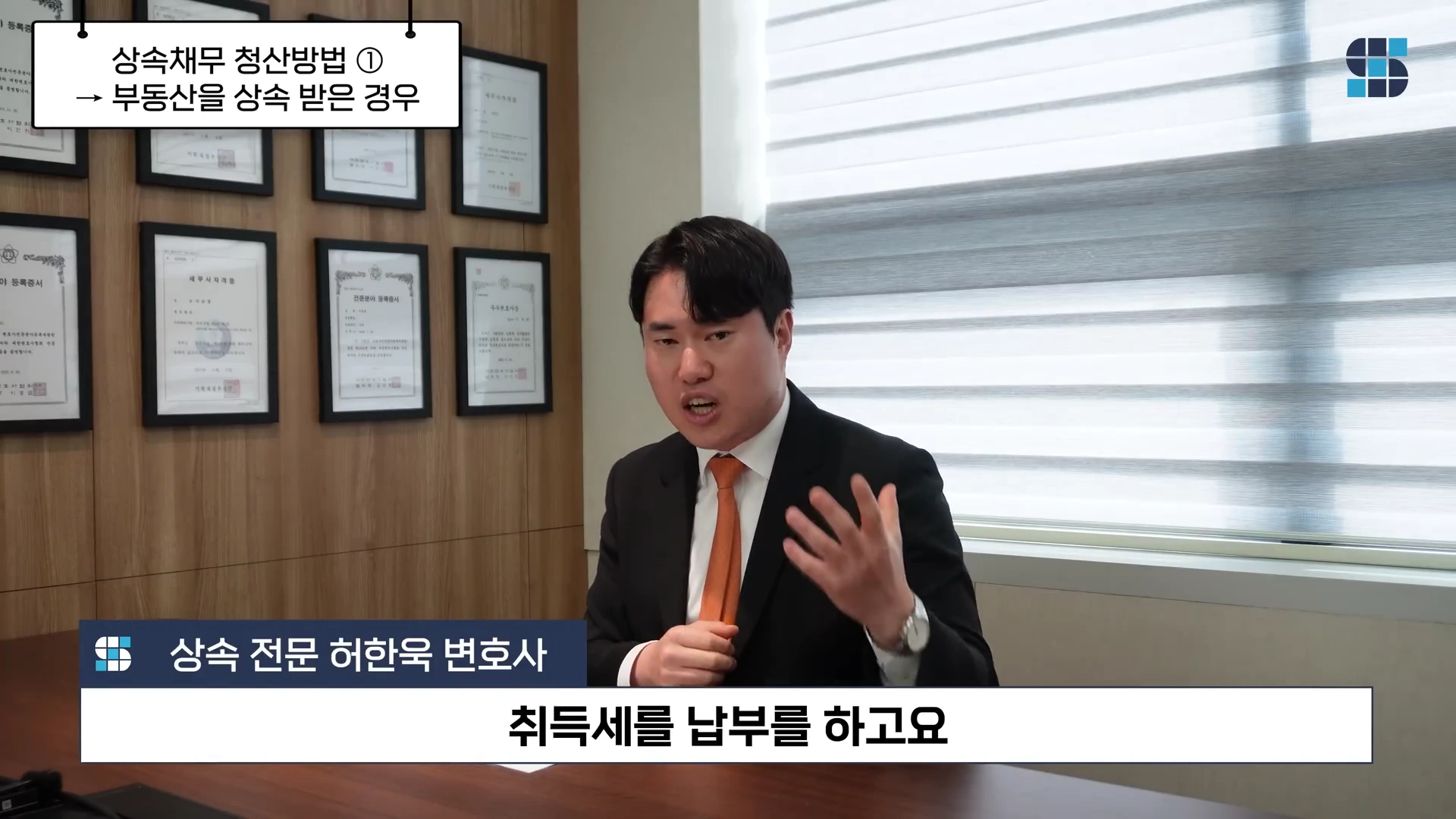 정장을 입은 남성이 사무실 책상에 앉아 손짓하며 설명하고 있고 화면 상단에는 '상속채무 청산방법 1. 부동산을 상속받은 경우'라는 자막이, 하단에는 '취득세를 납부를 하고요'라는 자막이 표시된 영상 화면.