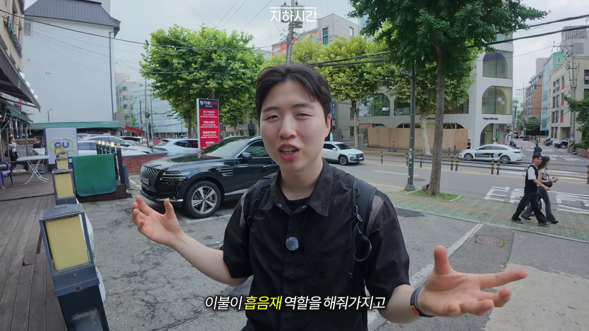 거리에서 검은색 셔츠를 입은 젊은 남성이 양손을 펼치며 이야기하고 있고, 배경에는 도로와 건물, 차량이 보입니다.