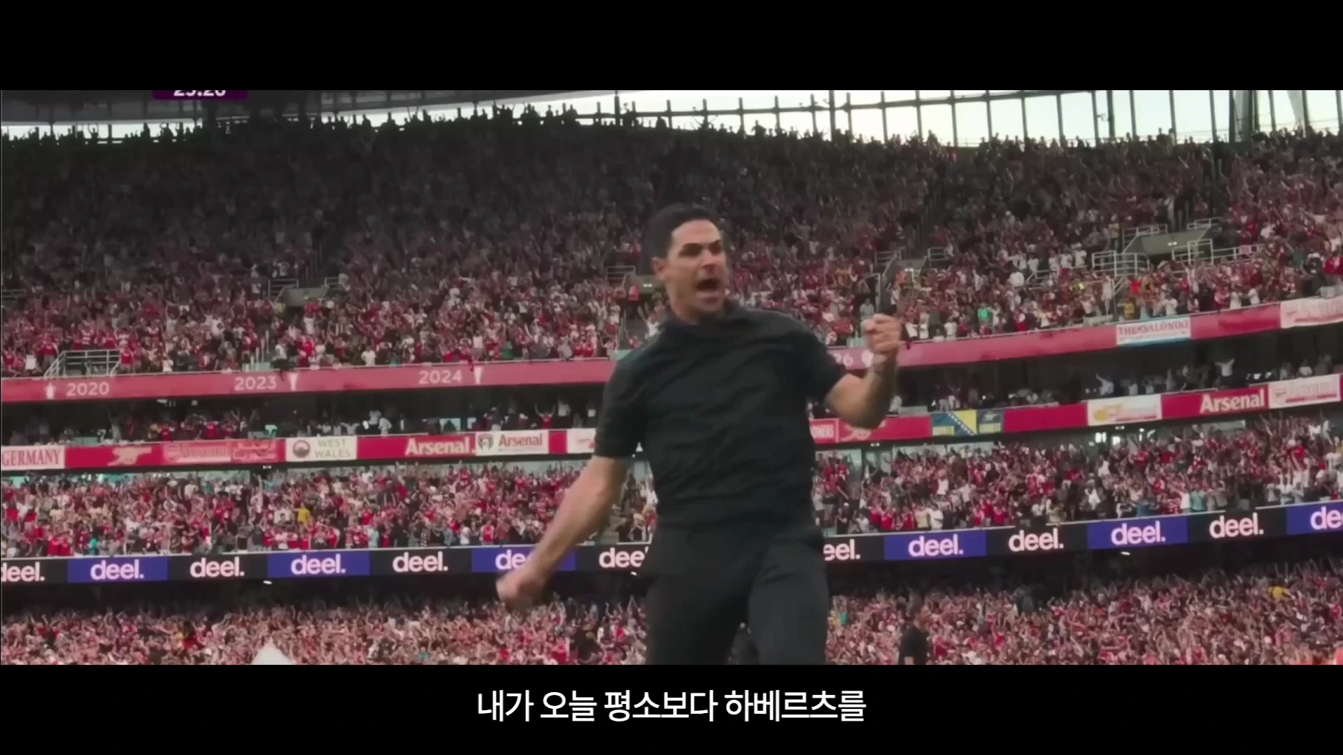 축구 경기장에서 검은 옷을 입은 감독이 열정적으로 주먹을 불끈 쥐며 환호하고 있는 장면