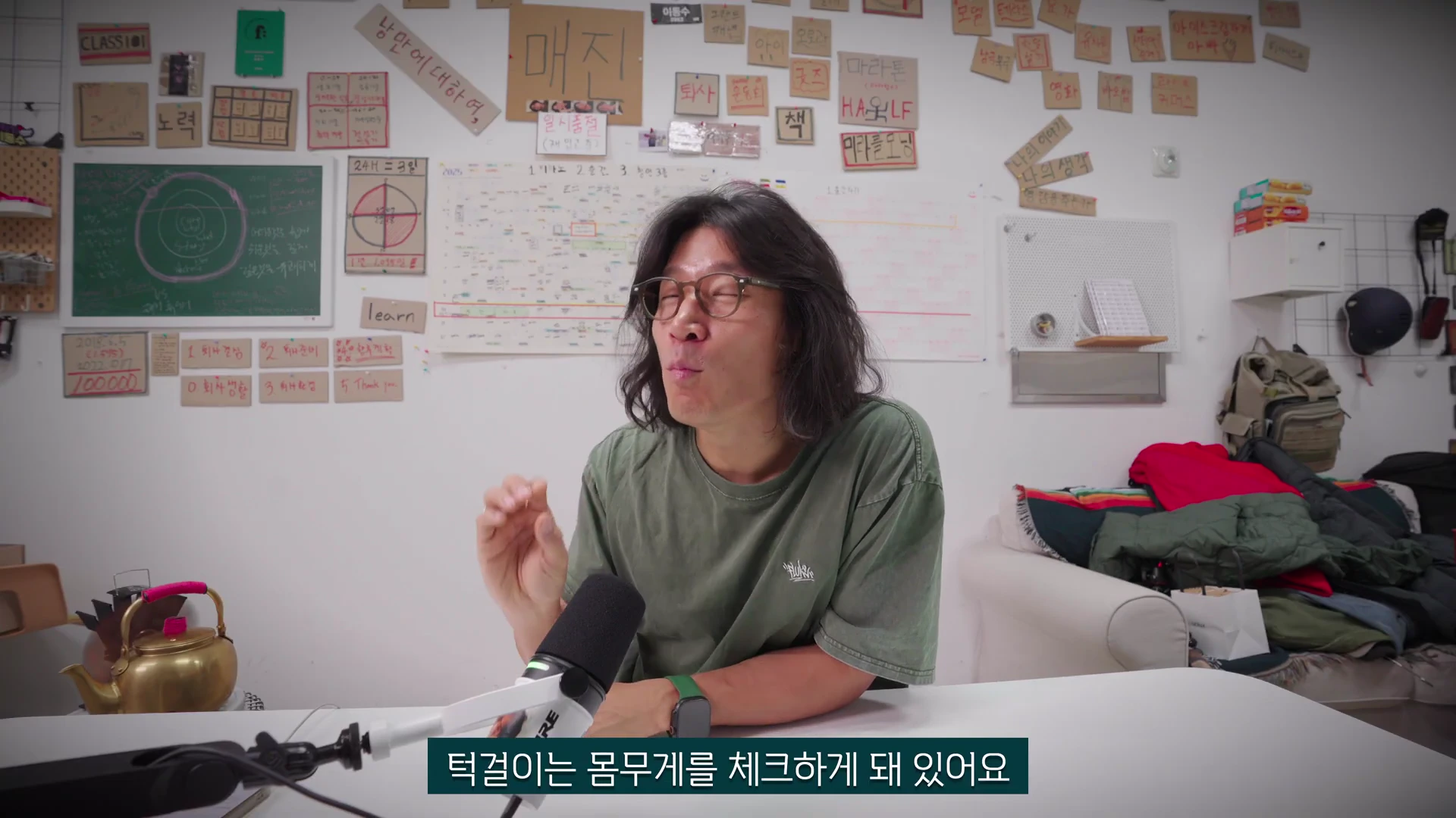 안경을 쓴 긴 머리의 남성이 책상 앞에 앉아 마이크에 대고 이야기하고 있으며, 뒤쪽 벽면에는 다양한 메모와 포스트잇이 붙어 있습니다.