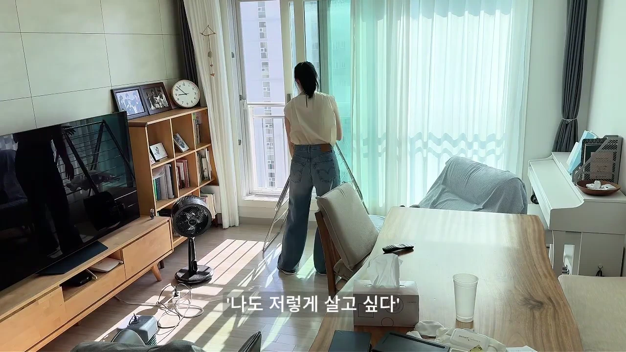 햇살이 들어오는 거실 창가에 서 있는 여성과 깔끔하게 정돈된 거실 풍경이 담긴 영상 캡처 화면입니다.