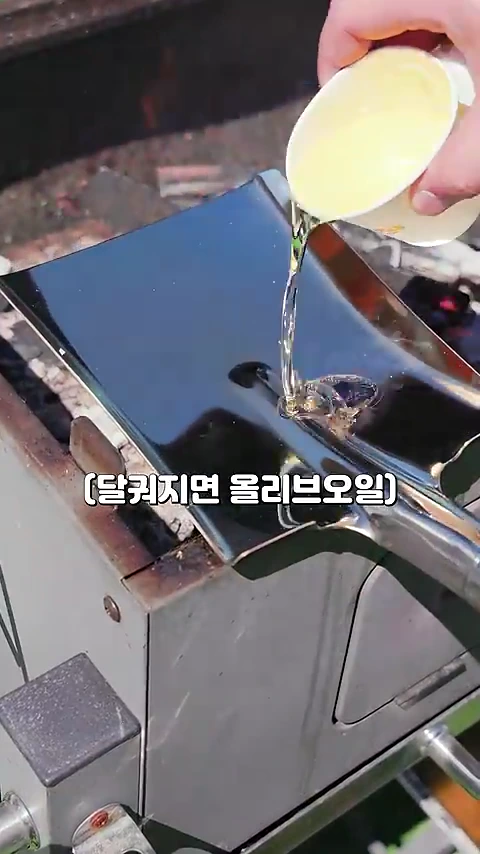 숯불 위에 달궈진 조리용 삽 위에 종이컵에서 올리브 오일을 붓는 장면이 담긴 영상 캡처 화면입니다.