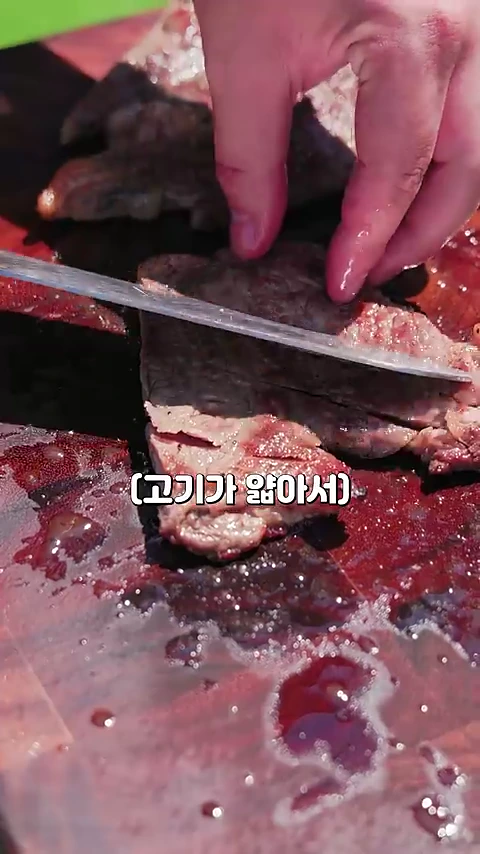 나무 도마 위에서 익힌 스테이크를 칼로 썰고 있는 손 모습과 화면 중앙에 (고기가 얇아서)라는 자막이 표시된 근접 촬영 영상 프레임