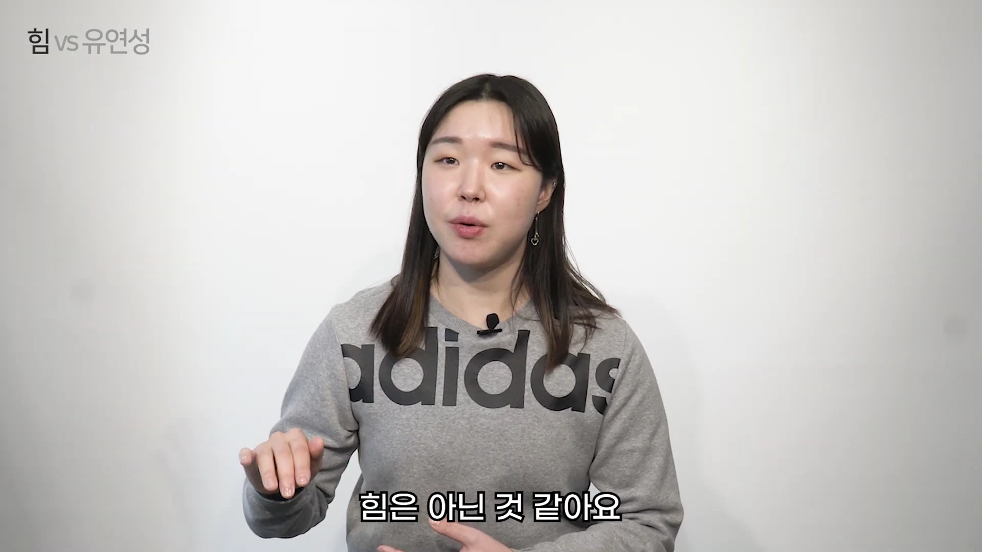 흰 벽을 배경으로 회색 아디다스 맨투맨을 입은 여성이 카메라를 정면으로 보며 이야기하고 있고, 화면 왼쪽 상단에는 '힘 vs 유연성'이라는 자막이, 하단에는 '힘은 아닌 것 같아요'라는 자막이 표시되어 있습니다.