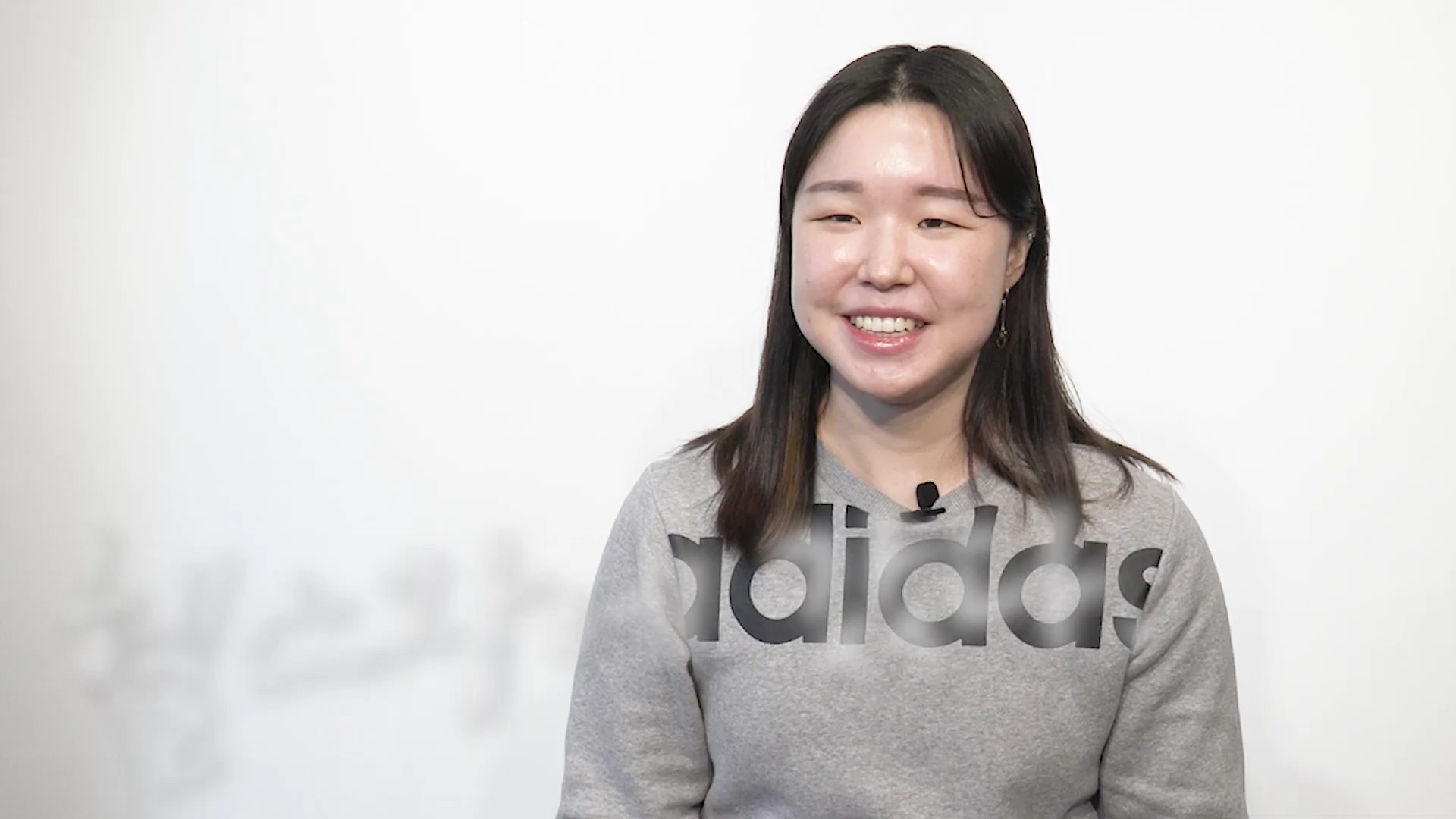 흰색 배경 앞에 회색 아디다스 맨투맨을 입은 여성이 밝게 웃으며 앉아 있는 상반신 모습입니다.