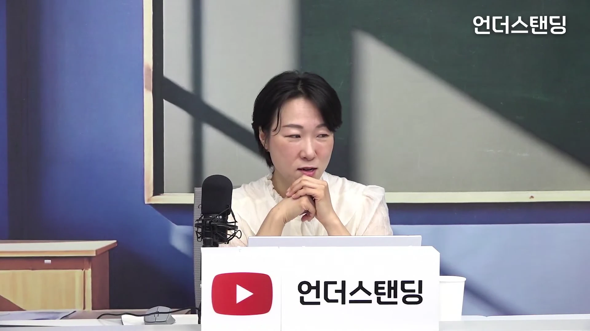 단발머리의 여성 출연자가 스튜디오에서 마이크 앞에 앉아 두 손을 모으고 이야기를 나누고 있다.