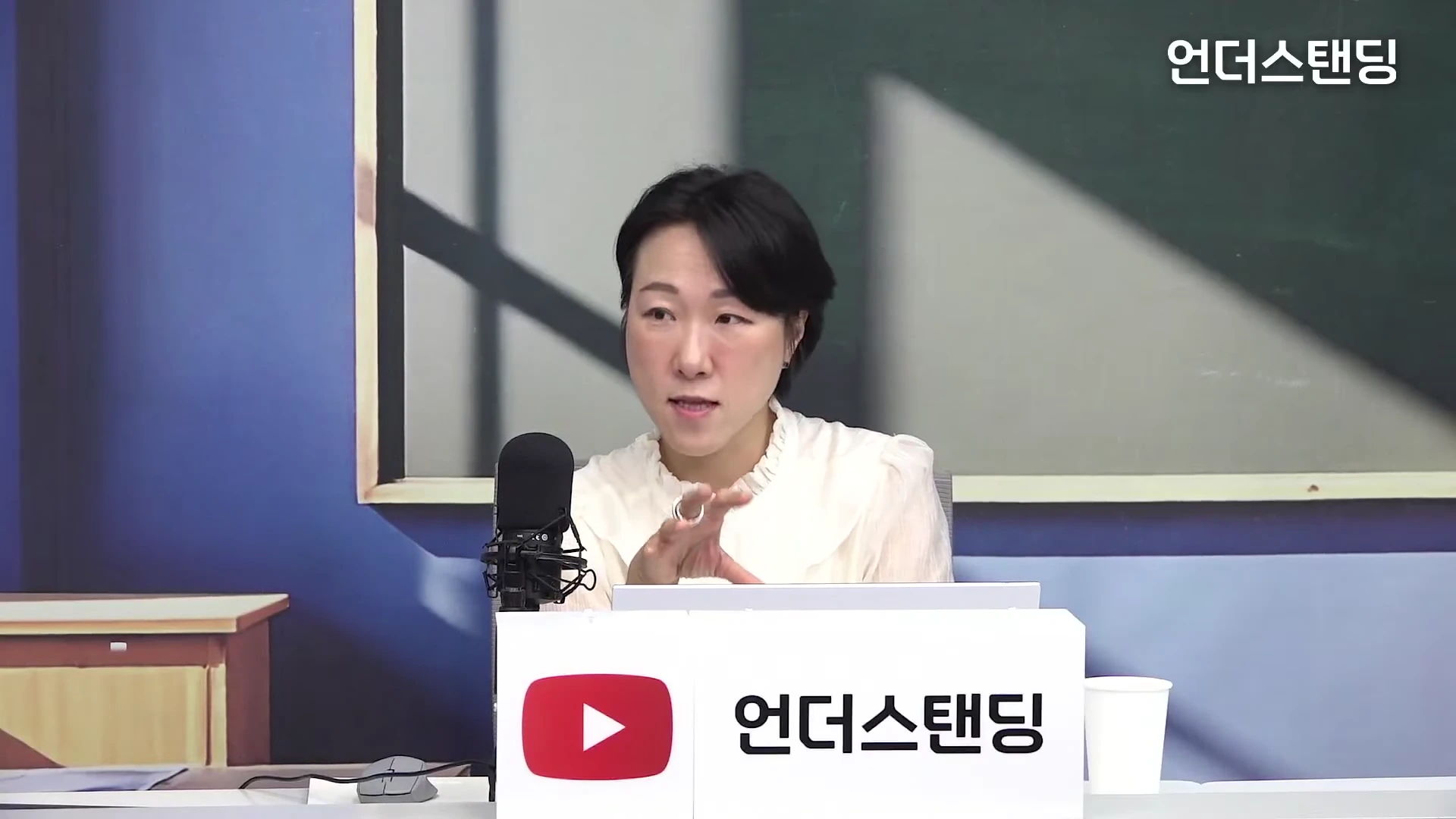 단발머리의 여성 출연자가 흰색 블라우스를 입고 스튜디오의 책상 앞에 앉아 손짓하며 대화하는 모습입니다.