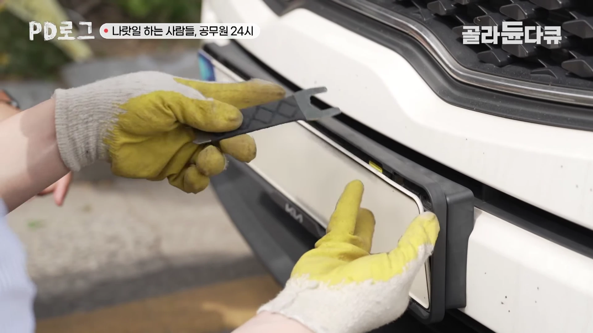 노란색 목장갑을 낀 손이 차량의 앞 번호판을 도구로 탈거하는 모습이 담긴 근접 촬영 화면.