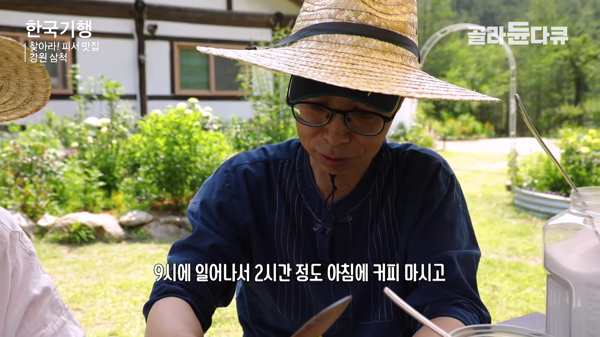 A person wearing a wide-brimmed straw hat and glasses is sitting outdoors in a sunny garden, with a blurred house and greenery in the background.