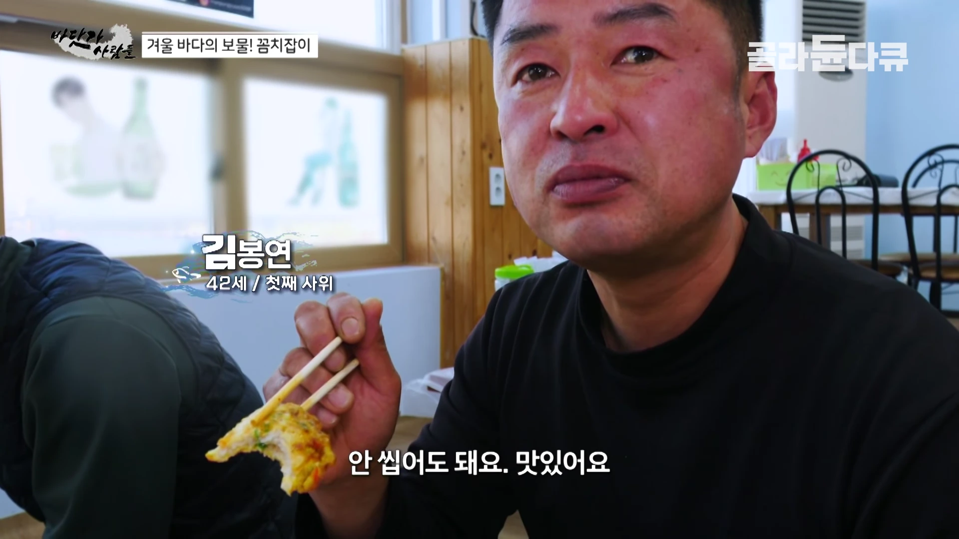 한 중년 남성이 식당에서 젓가락으로 음식을 들고 미소를 지으며 카메라를 바라보고 있는 모습