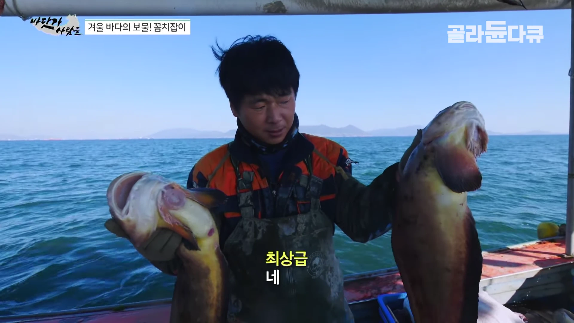 배 위에서 작업복을 입은 어부가 양손에 크고 길쭉한 꼼치를 한 마리씩 들고 있는 모습