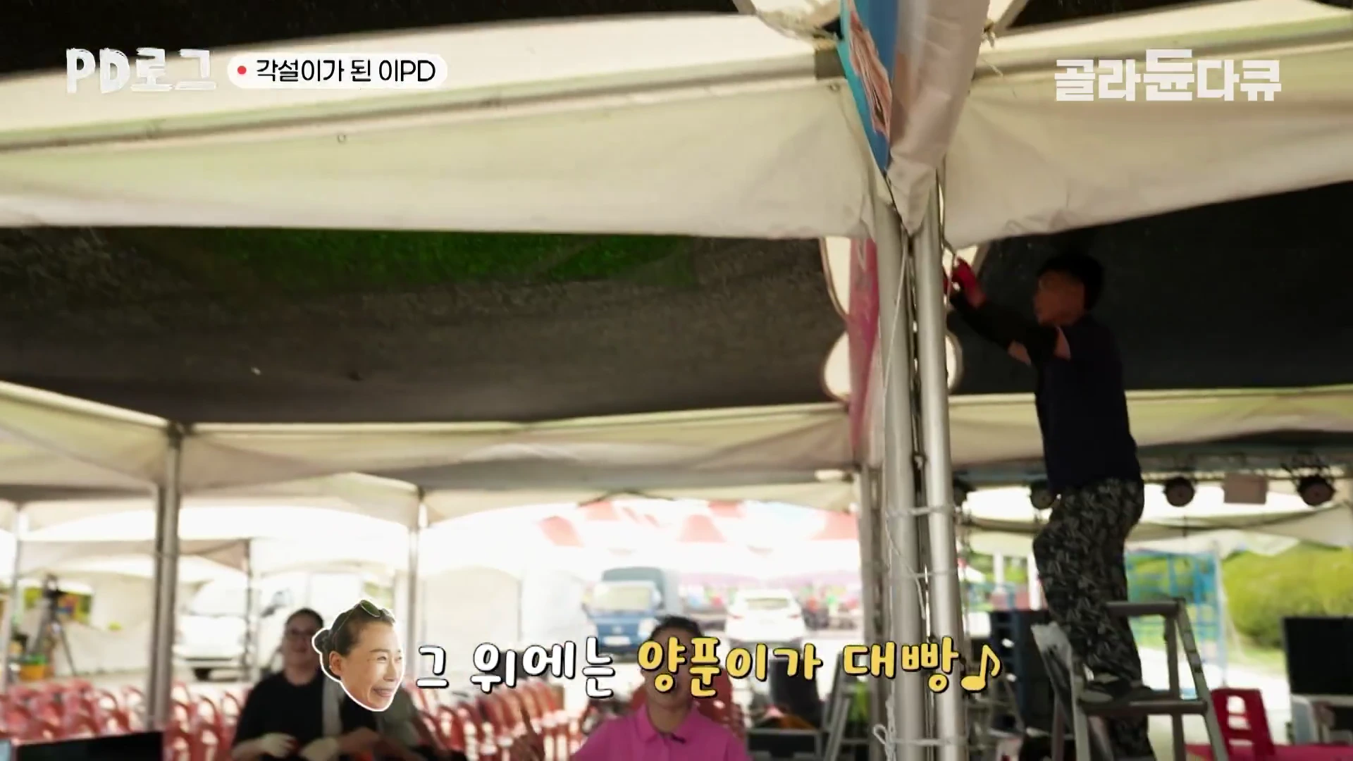 공연장 천막 기둥에 올라가 현수막을 고정하고 있는 사람과 아래에서 이를 지켜보는 사람들 모습