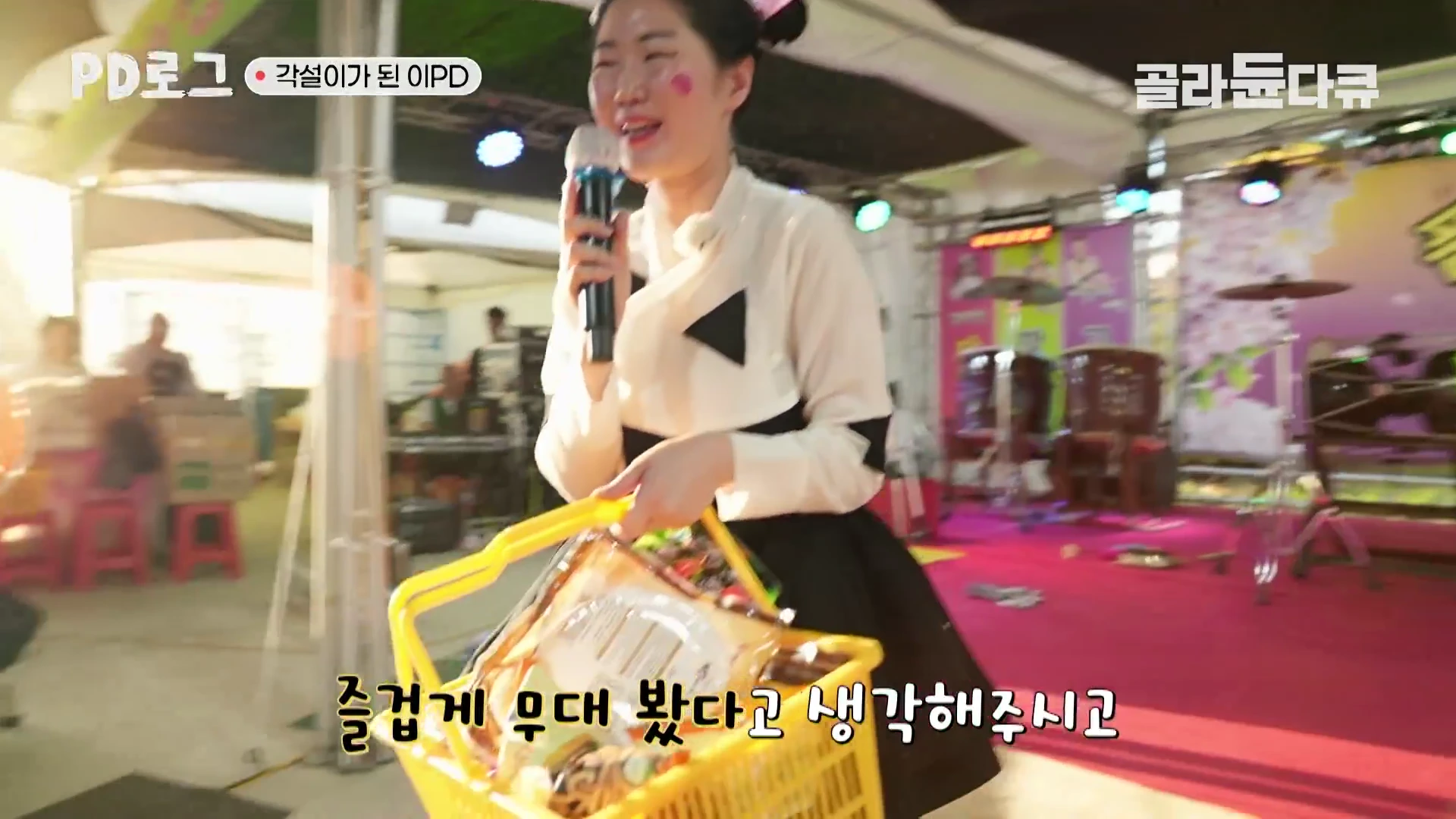 한 여성이 야외 무대에서 마이크를 잡고 노래하거나 말하며 노란색 바구니를 들고 관객석을 향해 걷고 있다.