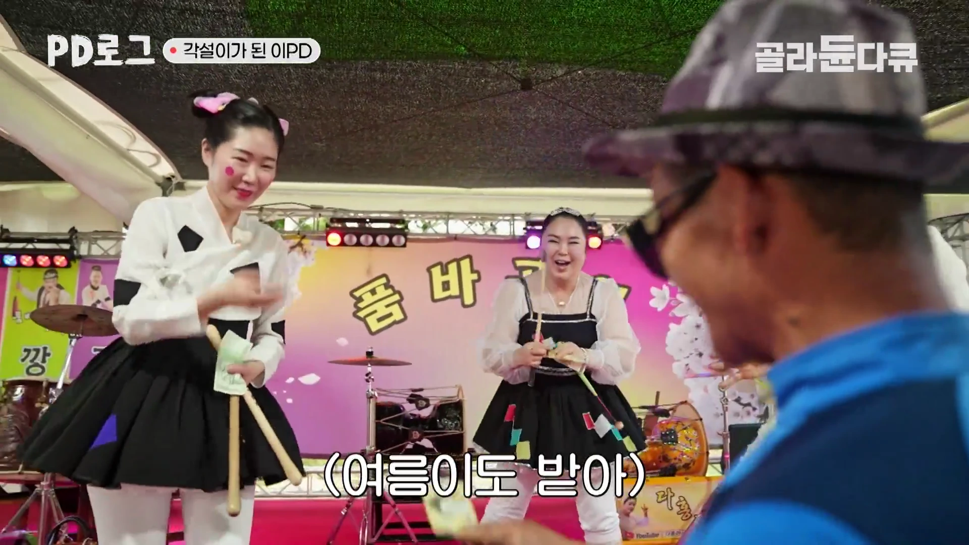 장터 무대 위에서 각설이 의상을 입은 두 명의 공연자가 관객으로부터 팁을 받아 들고 환하게 웃고 있는 모습
