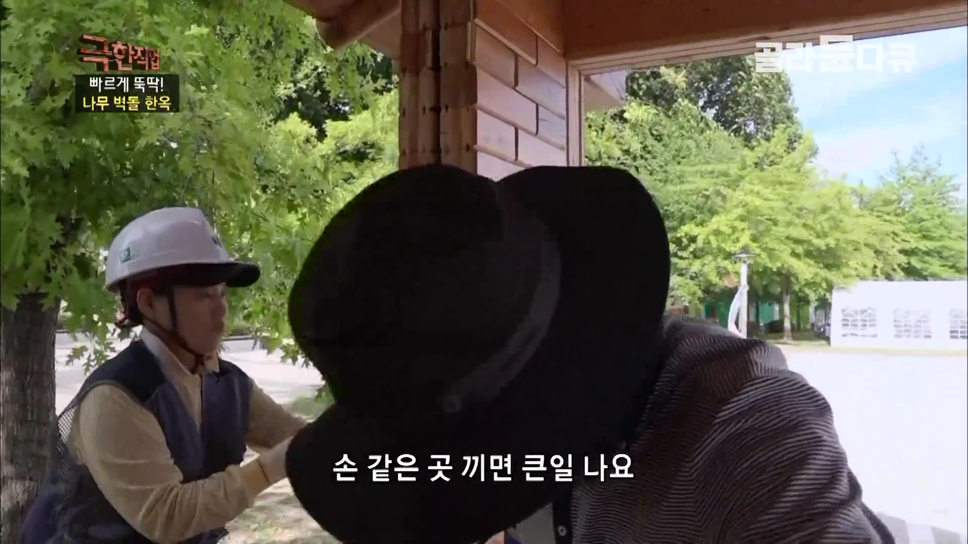 공사 현장에서 안전모를 쓴 작업자가 목재 벽체를 기둥 사이에 끼워 맞추고 있고, 그 앞에는 검은색 모자를 쓴 작업자가 주의 깊게 지켜보고 있습니다.