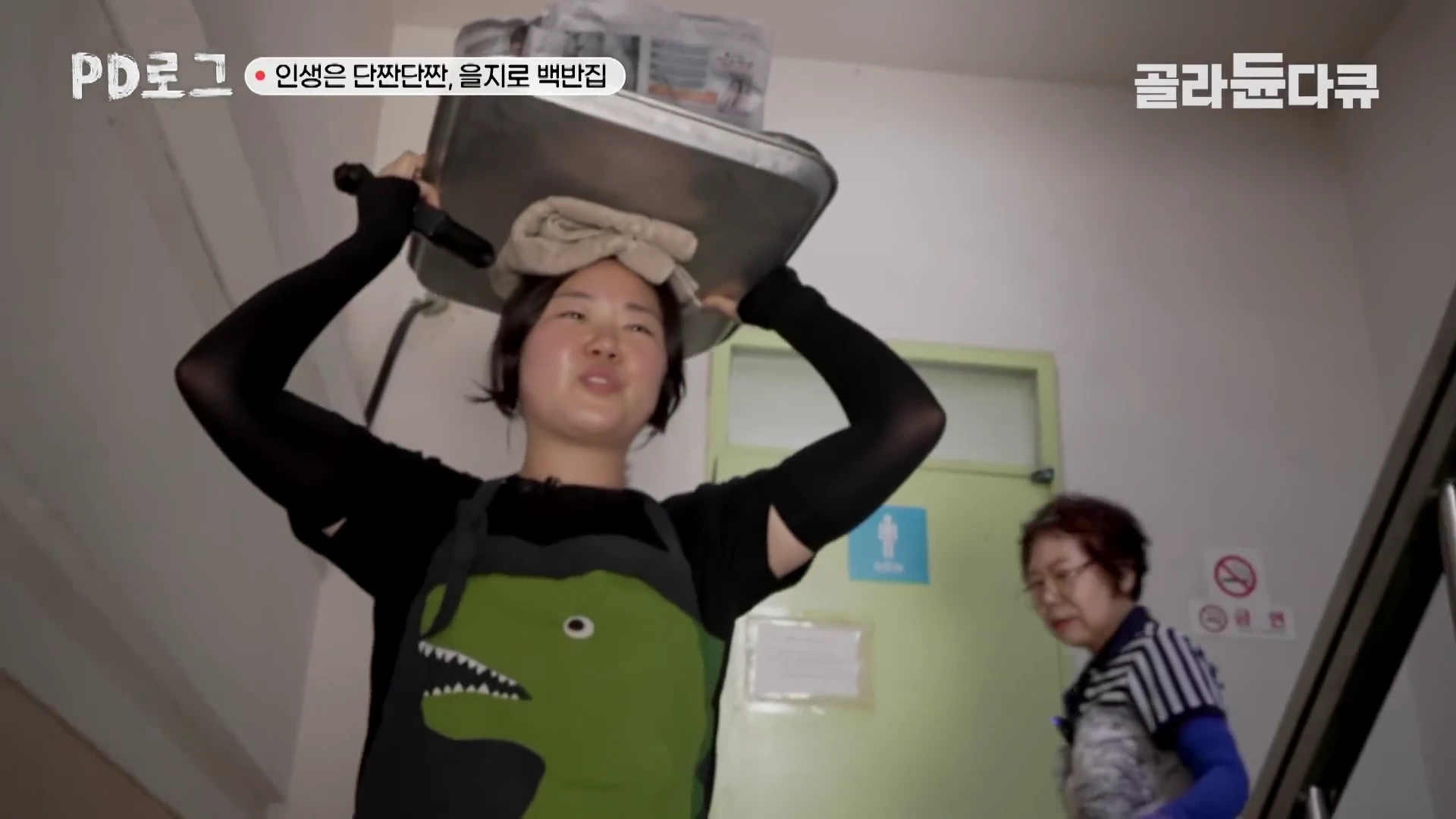 앞치마를 두른 젊은 여성과 뒤따르는 중년 여성이 좁은 계단을 오르며, 젊은 여성은 머리에 큰 은쟁반을 이고 있다.