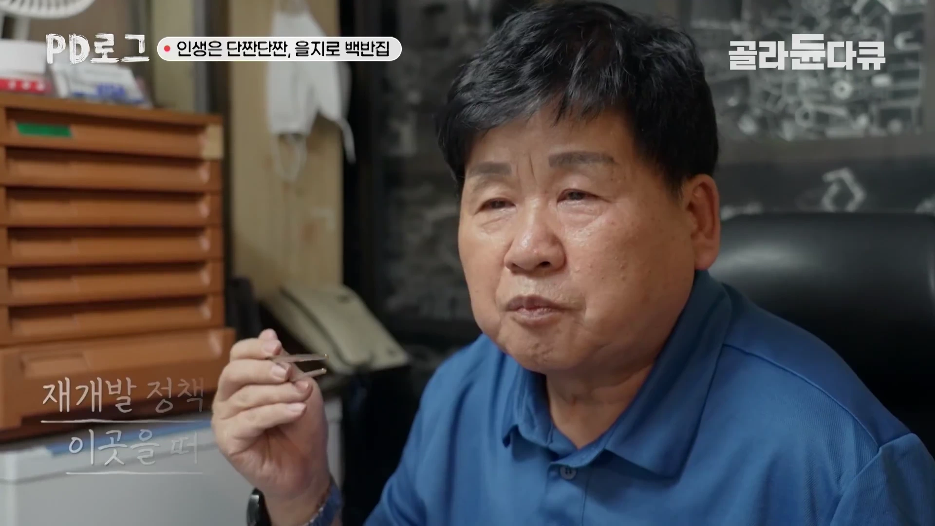 푸른색 셔츠를 입은 중년 남성이 실내 사무 공간에서 인터뷰하는 모습으로, 화면 상단과 하단에는 자막이 포함되어 있습니다.
