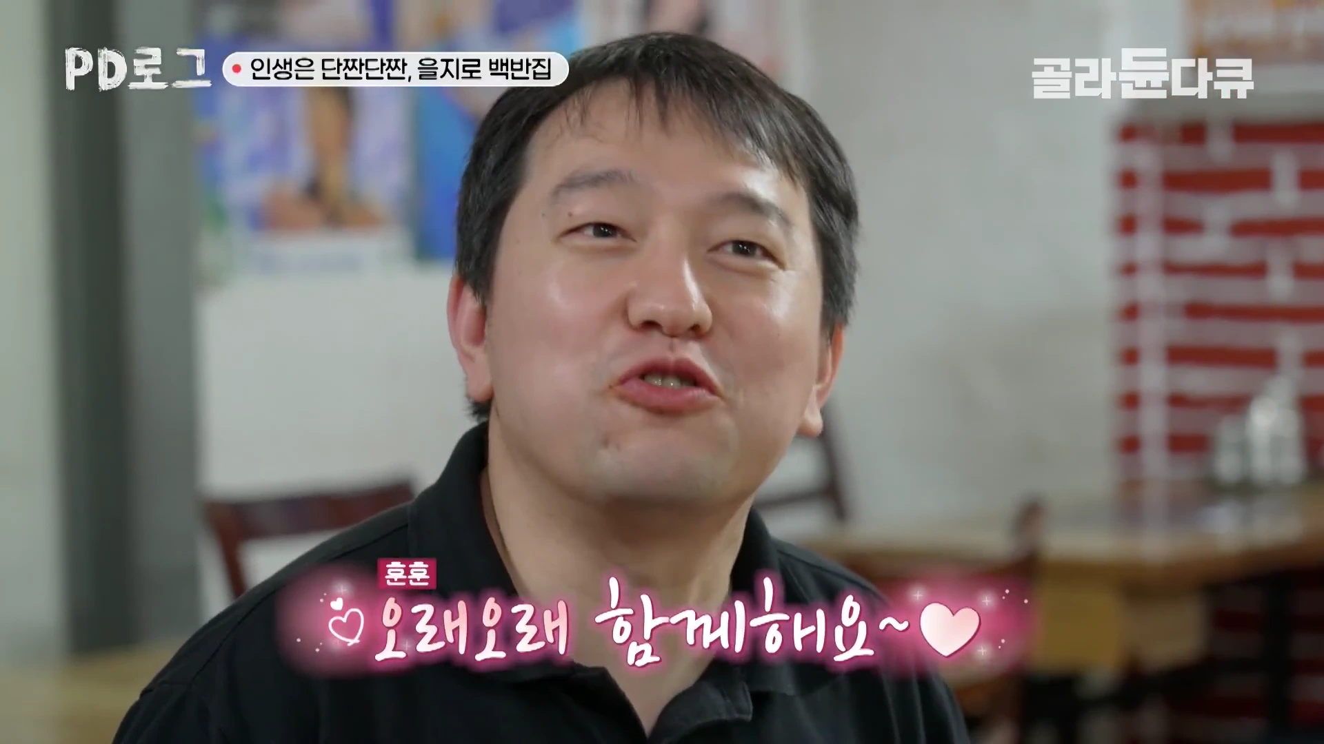 검은색 폴로 셔츠를 입은 중년 남성이 식당 내부에서 웃으며 말하고 있는 인터뷰 장면.