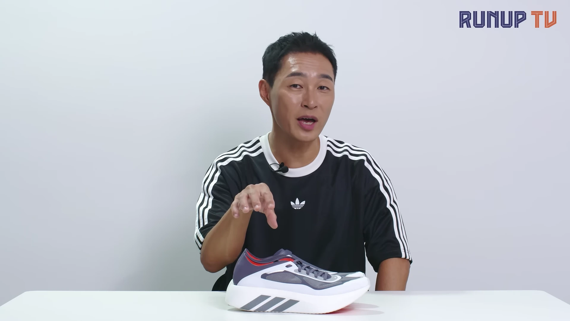 흰색 배경의 스튜디오에서 남성 진행자가 테이블 위에 놓인 운동화를 손으로 가리키며 설명하고 있다.