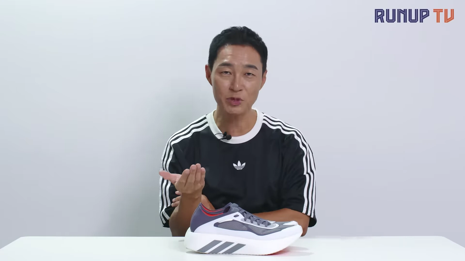 흰 배경의 스튜디오에서 한 남성이 아디다스 러닝화를 책상 위에 올려두고 손짓하며 이야기를 나누고 있다.