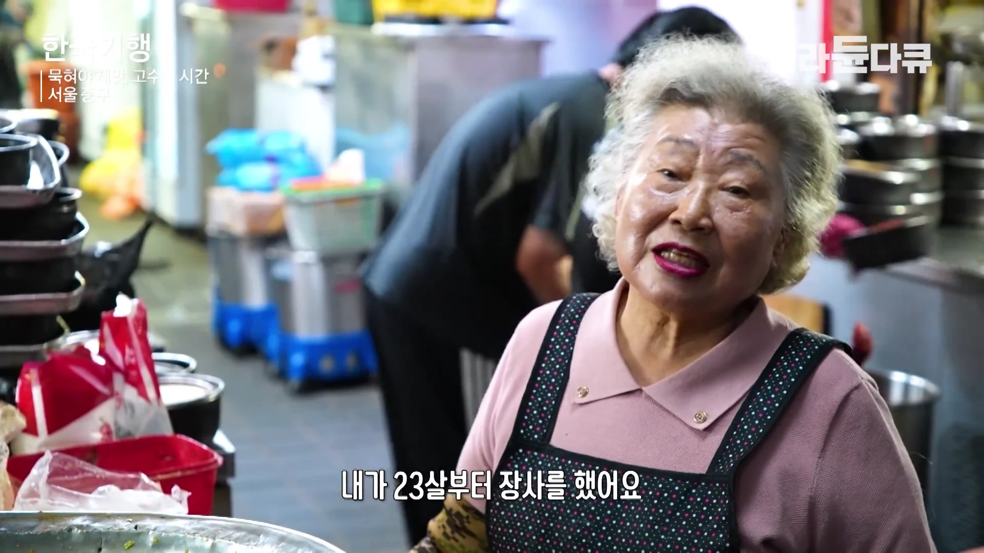 앞치마를 두른 은발의 여성 상인이 식당 주방에서 카메라를 바라보며 이야기를 나누고 있는 모습이다.