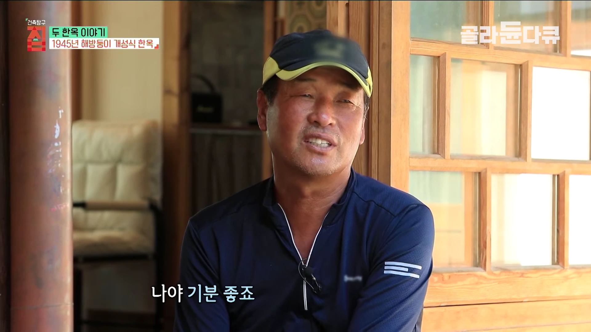 실내에서 야구 모자를 쓴 중년 남성이 인터뷰를 진행하고 있으며, 뒤로 나무 문과 창틀이 보인다.
