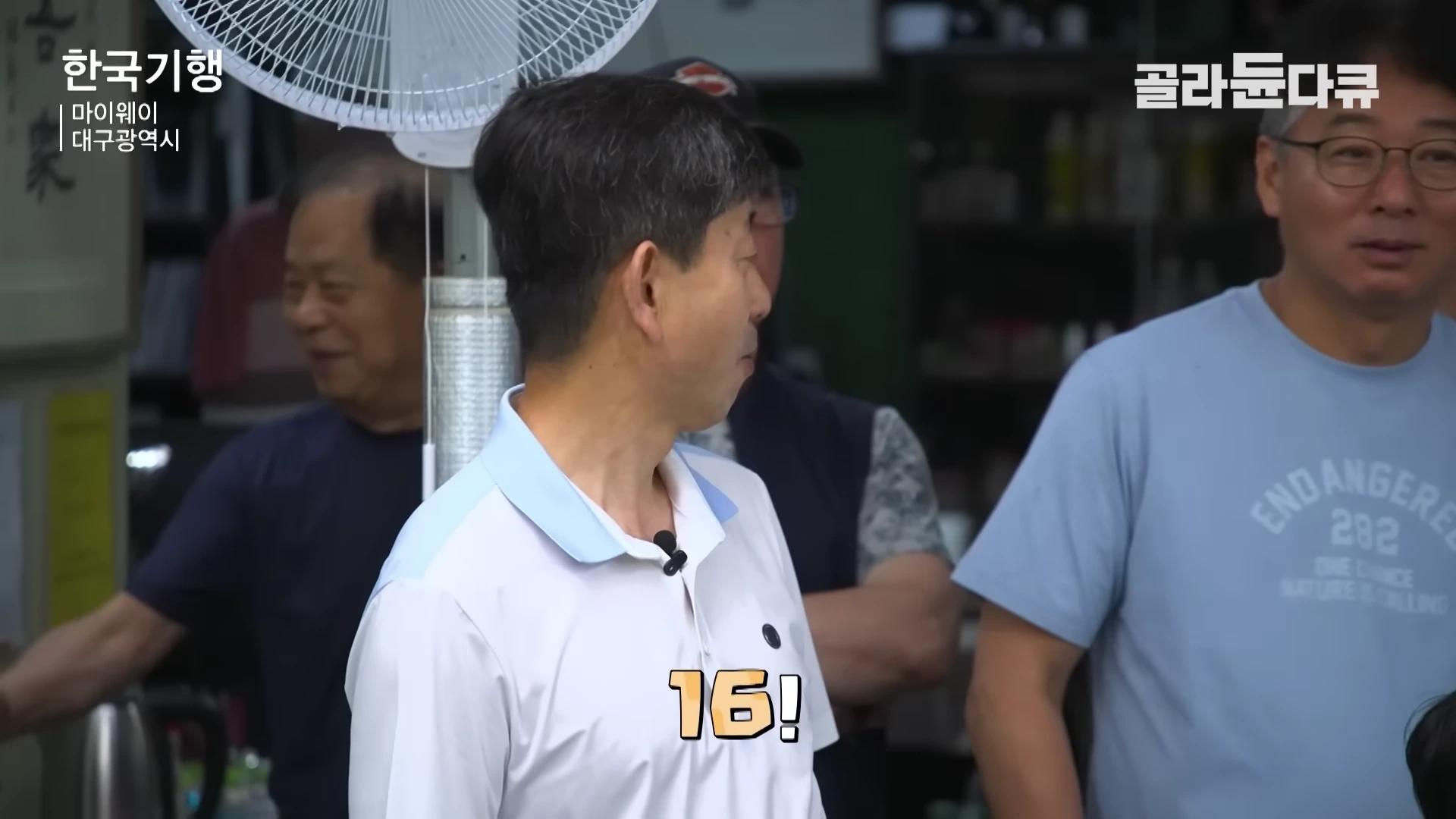실내에서 여러 사람이 모여 난초 경매를 진행하는 모습으로, 화면 중앙에는 숫자 16이 강조되어 표시되어 있습니다.
