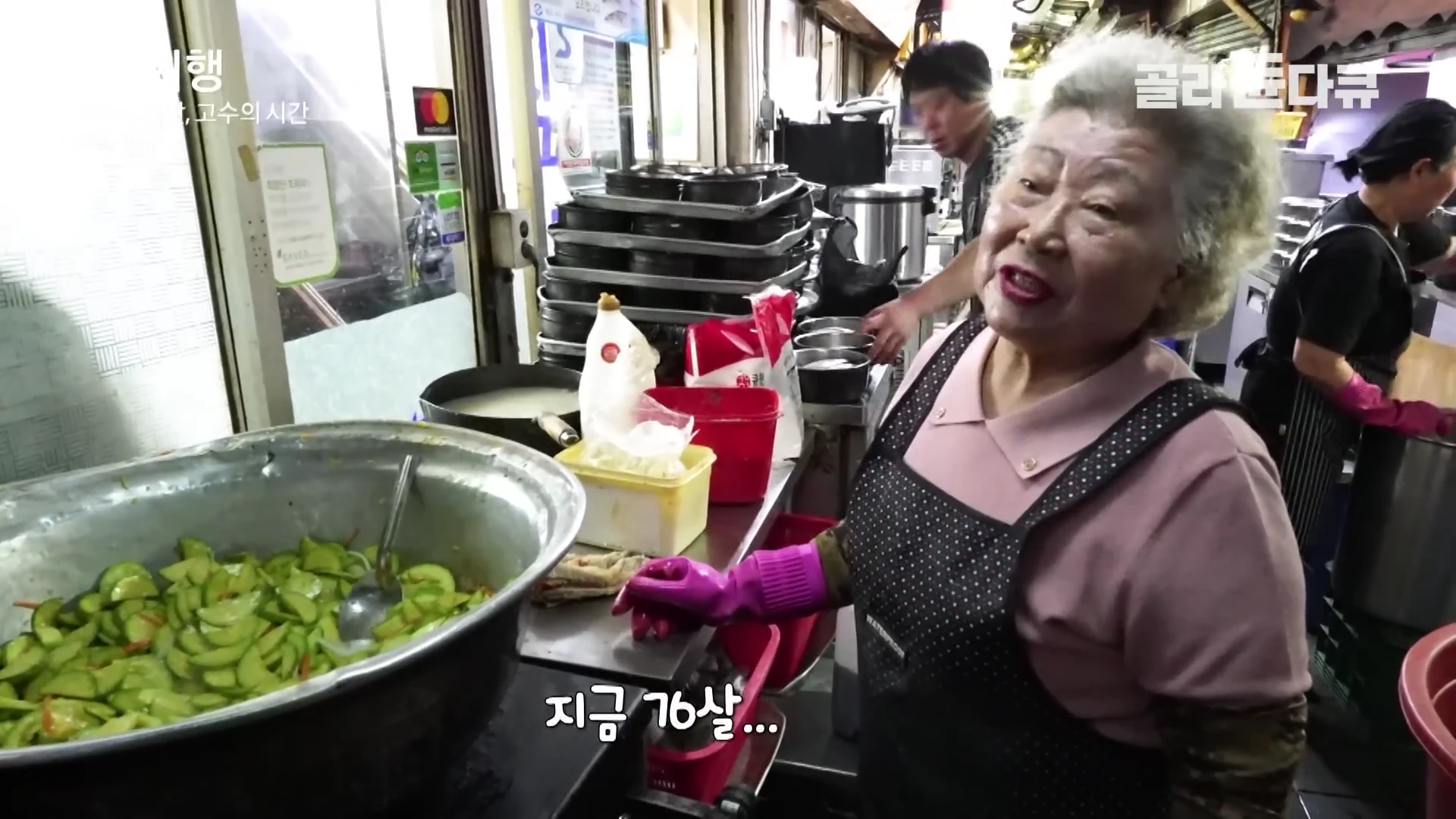 앞치마를 두른 백발의 할머니가 식당 주방에서 큰 솥에 담긴 애호박 볶음을 바라보며 이야기하고 있으며, 뒤편에는 다른 직원들이 바쁘게 움직이는 모습이 보입니다.