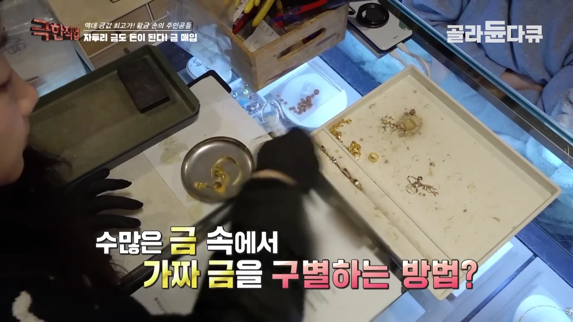 검은 장갑을 낀 손이 유리 진열대 위에 놓인 다양한 금 장신구와 부품들을 하나씩 분류하며 살피고 있는 모습