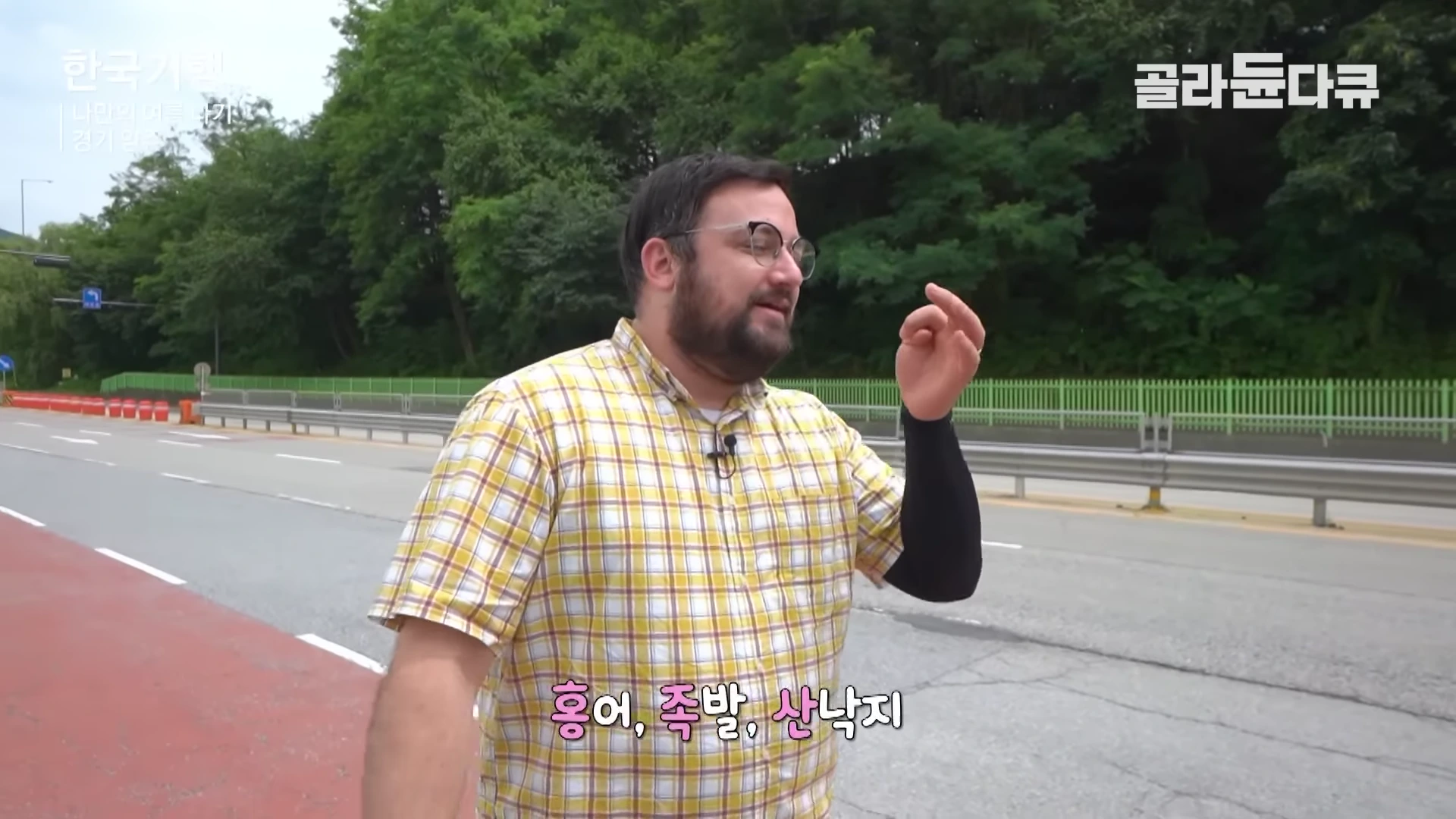 노란색 체크무늬 셔츠를 입고 안경을 쓴 외국인 남성이 도로 옆에서 손짓하며 이야기하고 있는 모습.