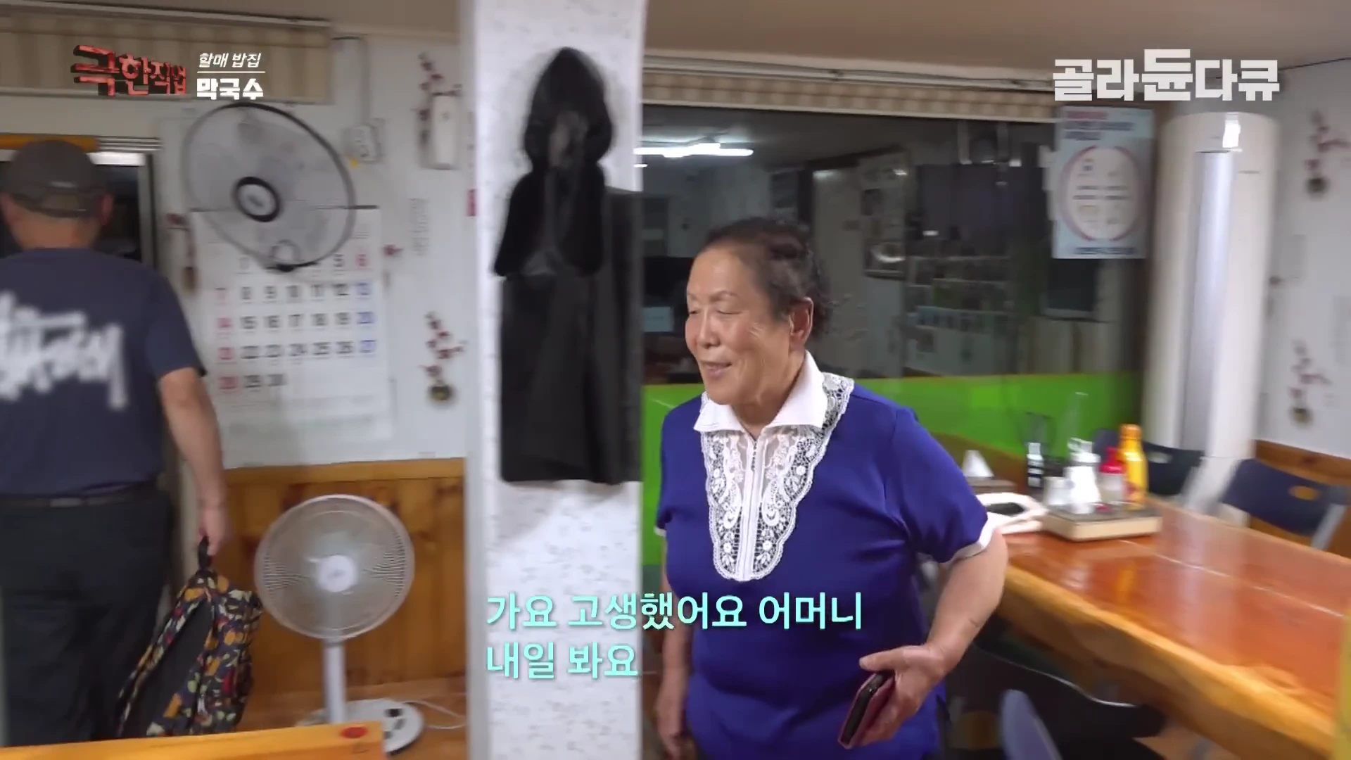 식당 내부에서 남색 상의를 입은 연세 지긋한 여성분이 밝게 웃으며 이동하고 있고, 그 뒤로 한 남성이 걸어 나가는 모습이 담긴 방송 화면이다.