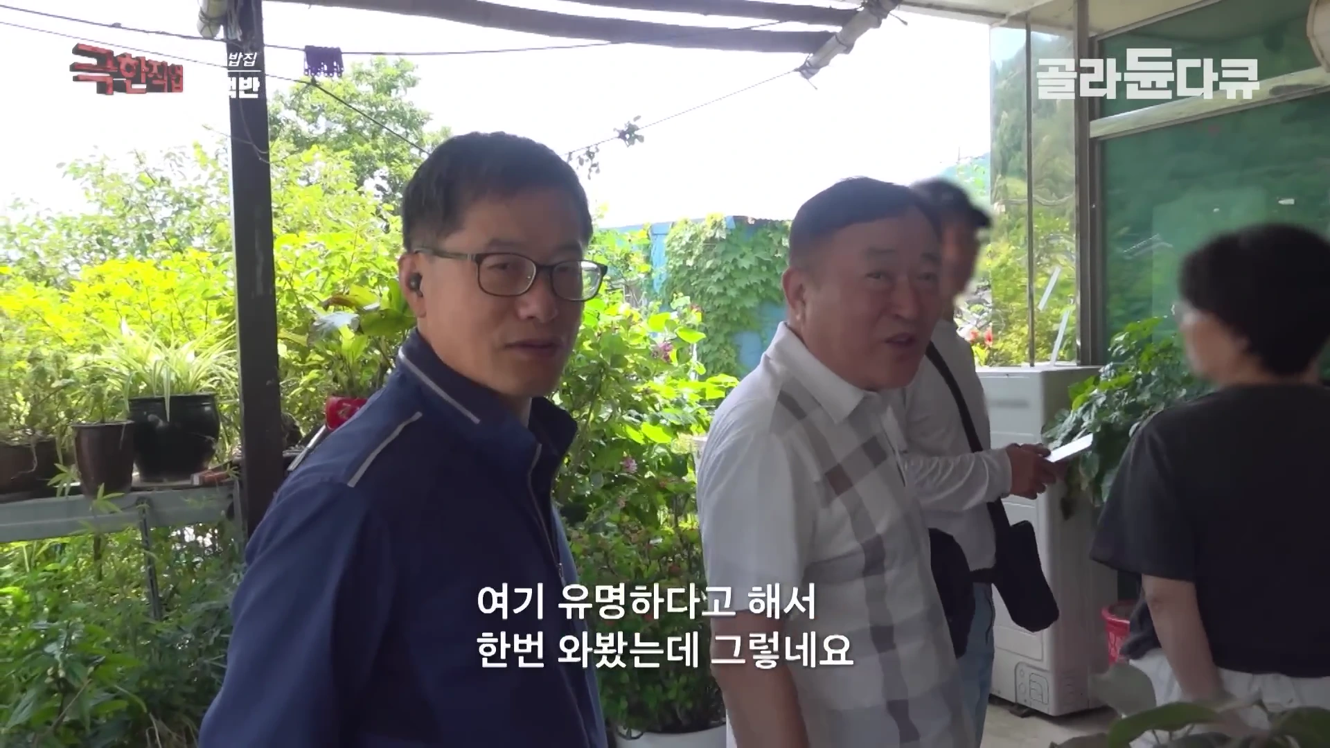 식당 입구에서 안경을 쓴 중년 남성이 카메라를 향해 이야기를 하고 있으며, 그 옆에는 흰색 체크무늬 셔츠를 입은 다른 남성이 함께 서 있다.