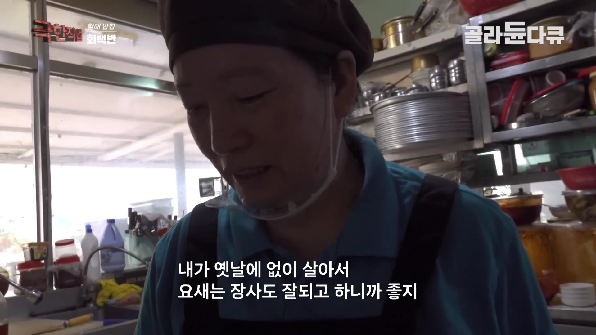 주방에서 일하는 중인 중년 여성의 모습이 담긴 화면으로, 여성은 푸른색 상의와 검은색 앞치마를 착용하고 있다.
