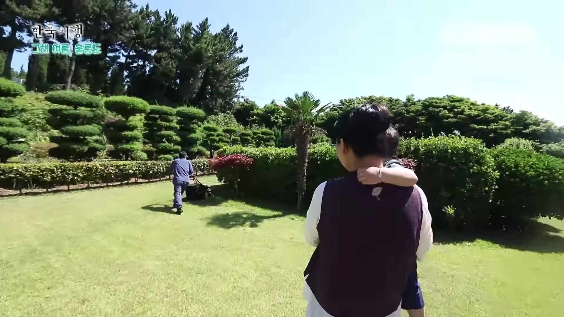 푸른 잔디가 깔린 넓은 정원에서 한 여성이 아이를 안고 있고, 그 앞쪽에서 한 남성이 잔디깎이 기계를 밀며 이동하고 있는 모습