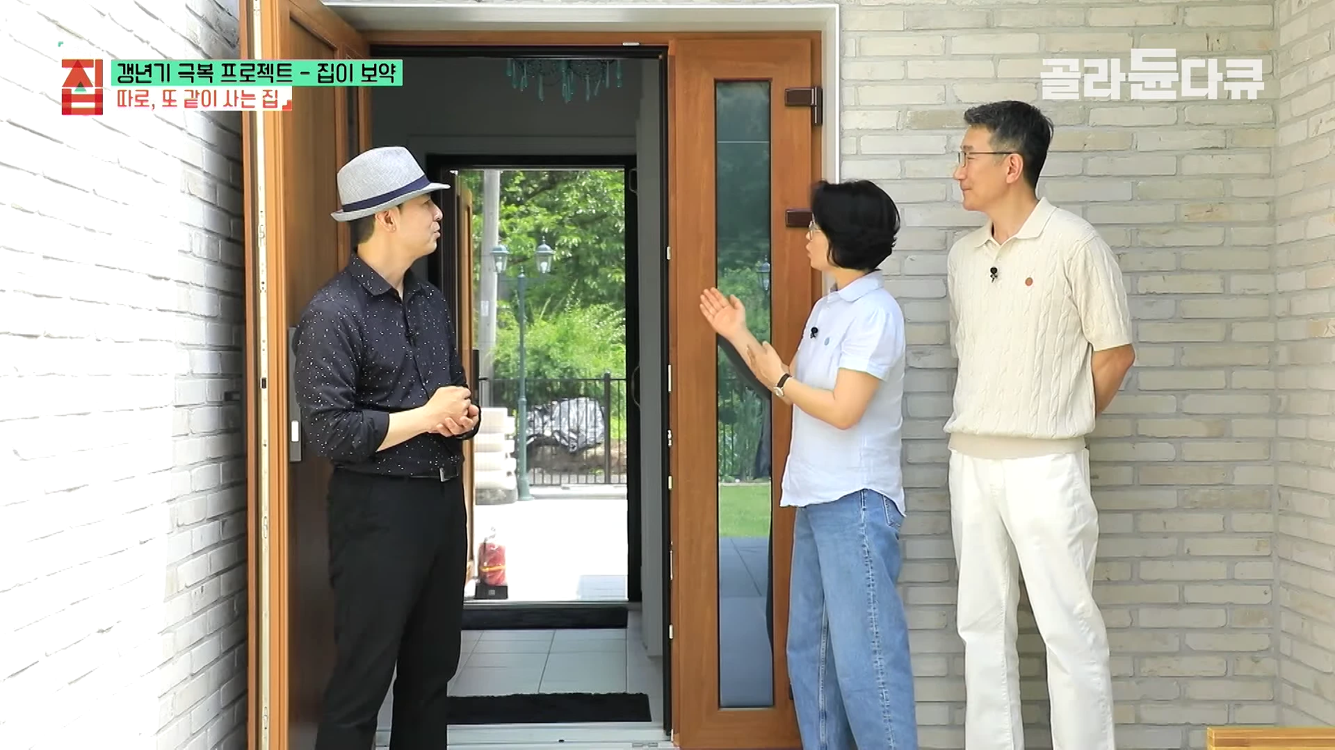 세 명의 사람이 밝은색 벽돌로 지어진 주택의 현관 앞에 서서 대화를 나누고 있으며, 열린 문 사이로 서로 다른 두 개의 중문이 보입니다.