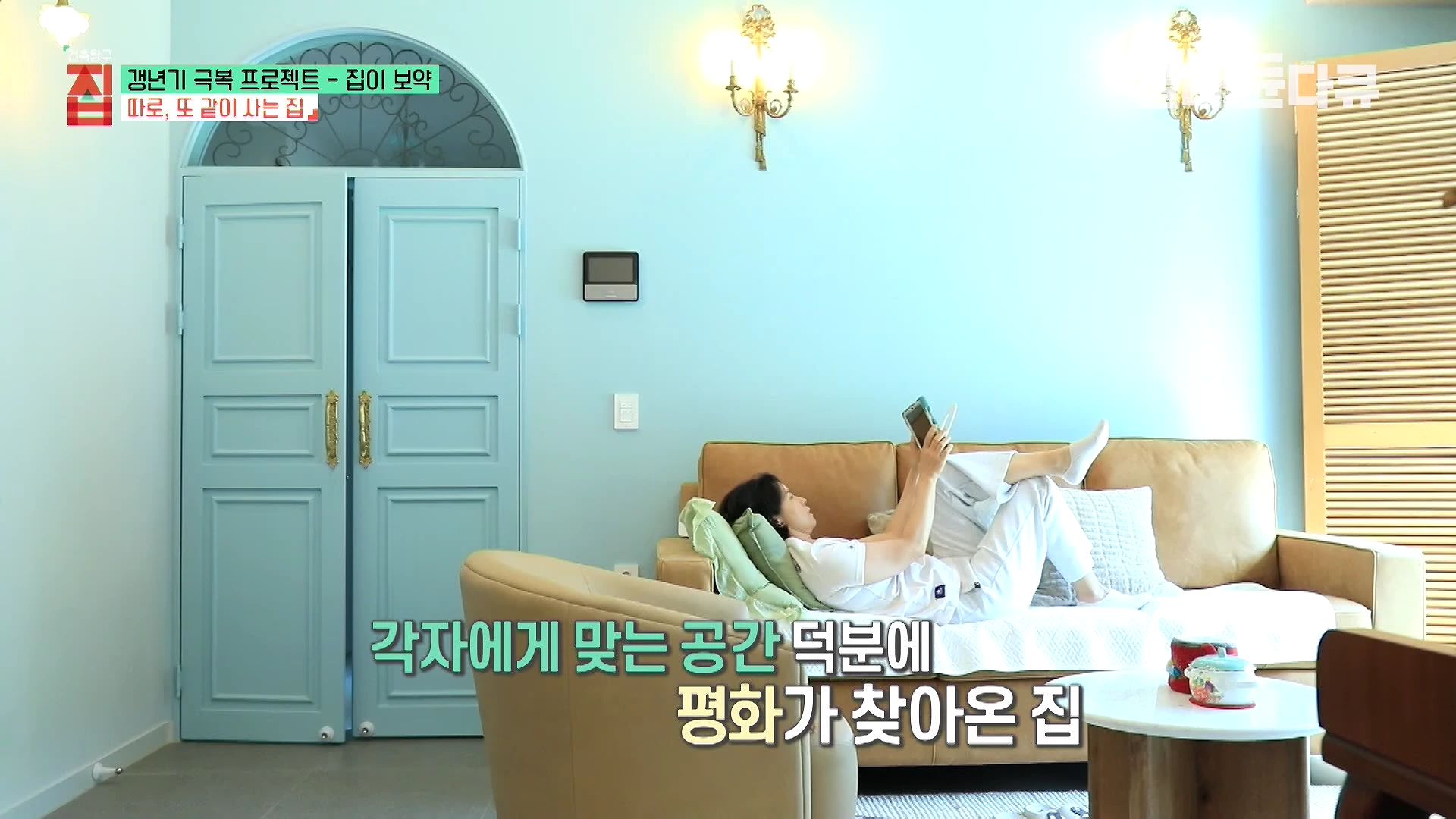 하늘색 벽과 아치형 문이 있는 거실에서 한 여성이 편안한 자세로 소파에 기대어 태블릿 PC를 보고 있다.
