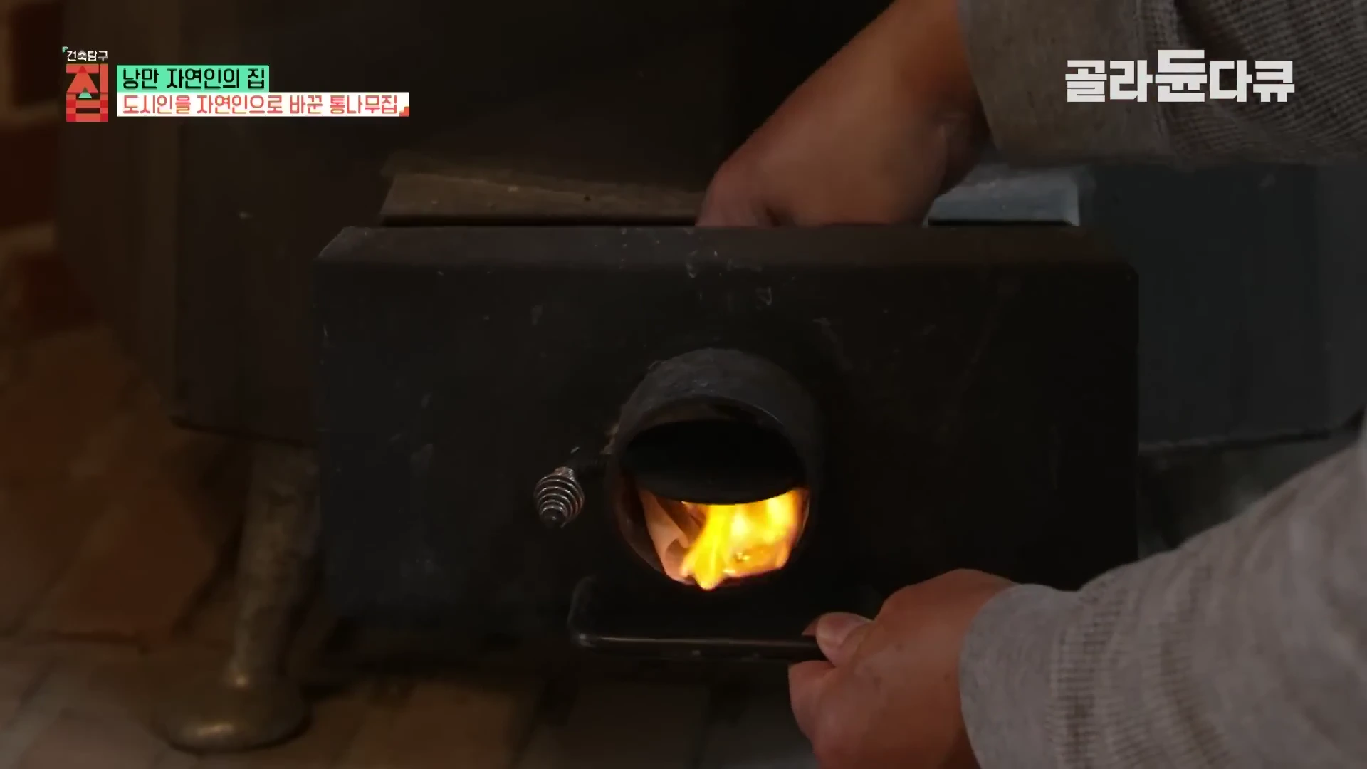 실내에서 사람이 검은색 화목 난로의 문을 열고 불을 지피고 있는 근접 촬영 영상 화면입니다.