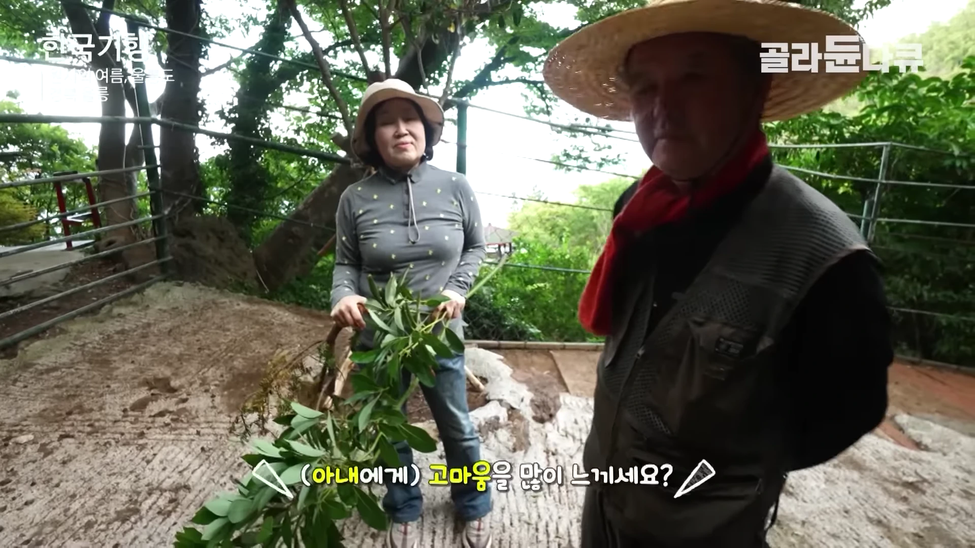 밀짚모자를 쓴 중년 남성과 벙거지 모자를 쓴 여성이 숲속 야외에서 나란히 서서 이야기를 나누는 모습