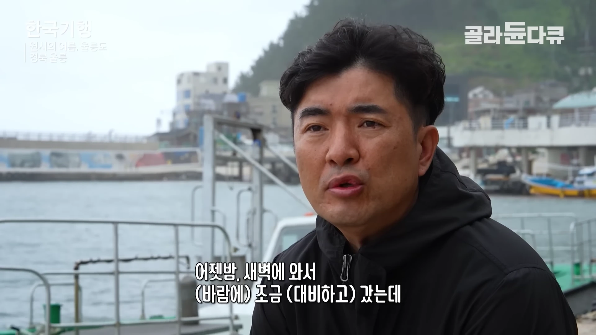 울릉도 항구 근처에서 검은색 재킷을 입은 한 중년 남성이 카메라를 보며 인터뷰하고 있다.