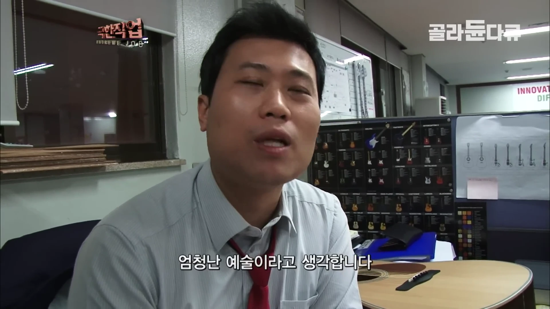사무실 책상 앞에 앉아 인터뷰 중인 남성, 배경에는 기타 모델이 나열된 게시판과 사무용품이 보임