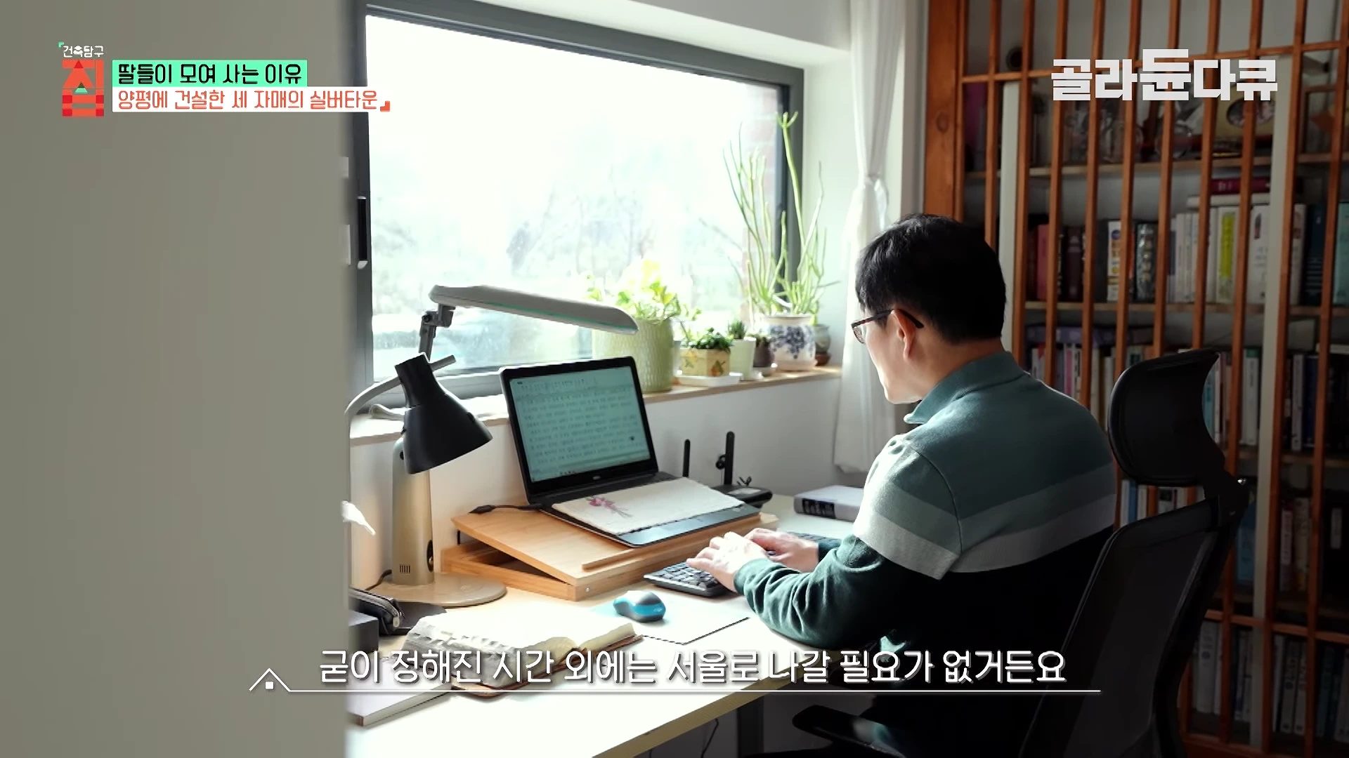 안경을 쓴 남성이 창가 책상에 앉아 노트북으로 글을 쓰고 있으며 옆에는 책장과 화분이 놓여 있다.