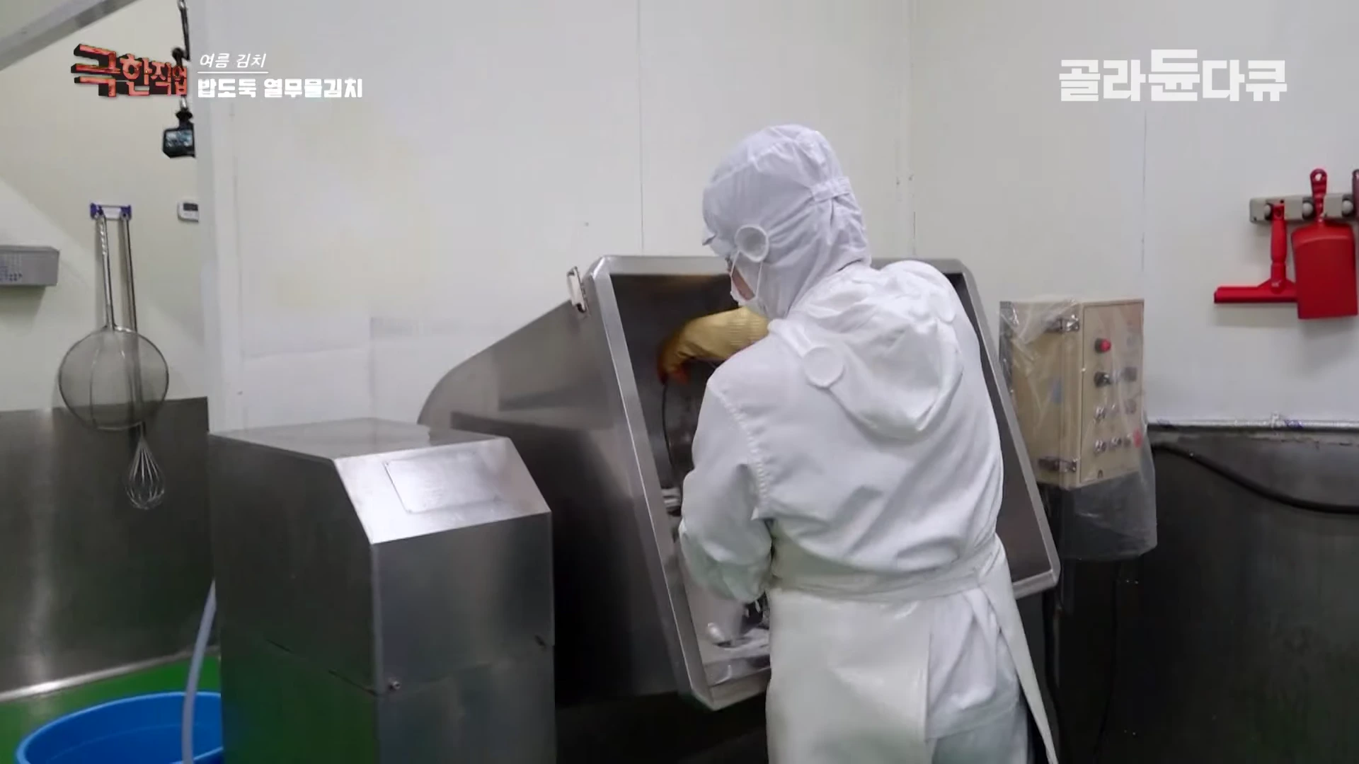 위생복과 위생모를 착용한 작업자가 금속 조리 설비 내부를 살피고 있는 김치 제조 시설의 모습