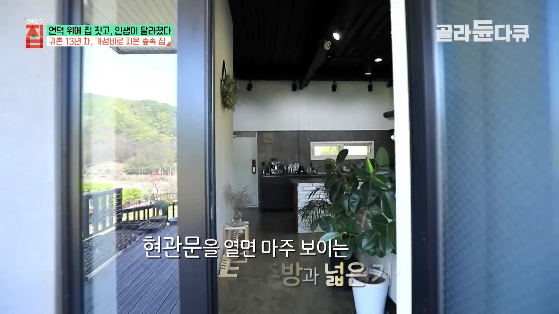 열린 현관문 너머로 신발을 신고 생활하는 넓은 거실과 주방이 보이며, 왼쪽 창문으로는 야외 데크와 산 풍경이 펼쳐져 있습니다.