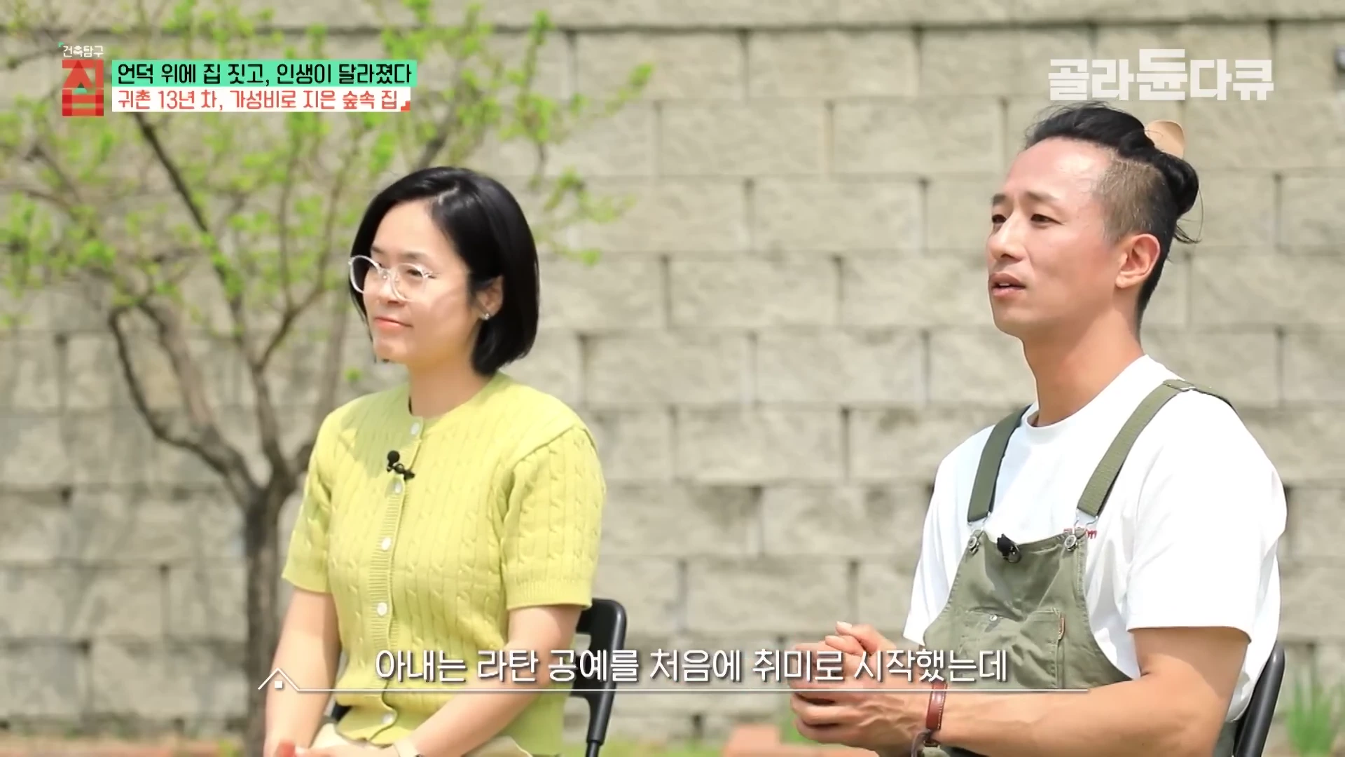실외에서 연두색 가디건을 입은 여성과 흰색 티셔츠에 멜빵 바지를 입은 남성이 나란히 앉아 인터뷰하는 모습