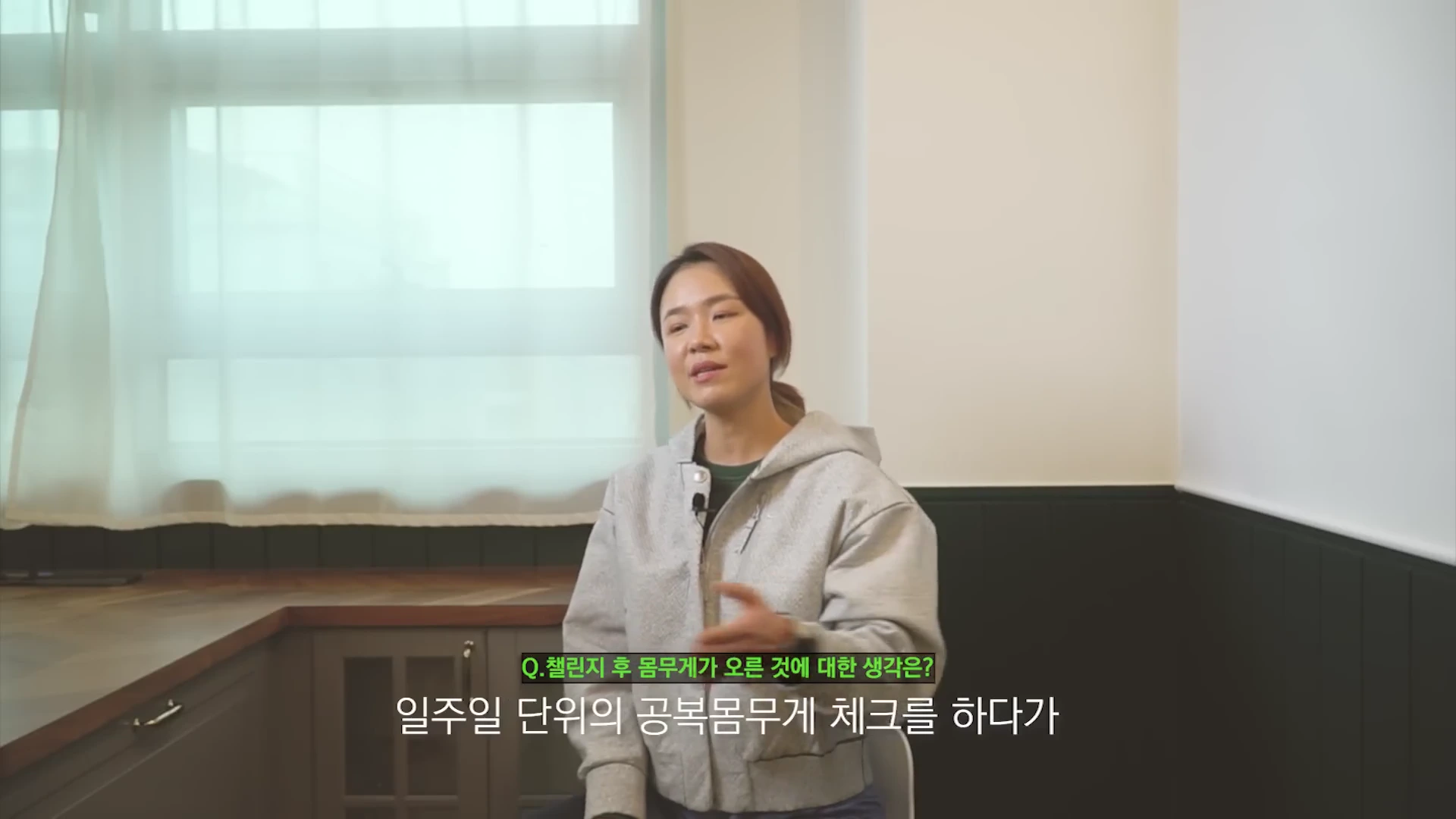 실내에서 회색 후드집업을 입은 여성이 앉아 인터뷰를 진행하고 있으며, 하단에는 질문 자막이 표시되어 있습니다.